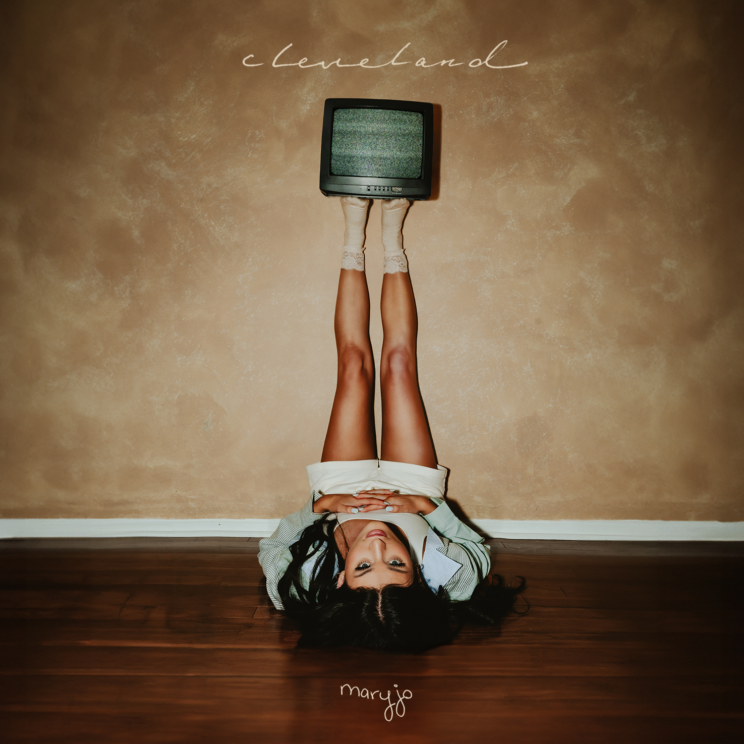 The cover art for Cleveland: maryjo lays on the floor with her legs propped up on the wall and a television balancing on her feet.